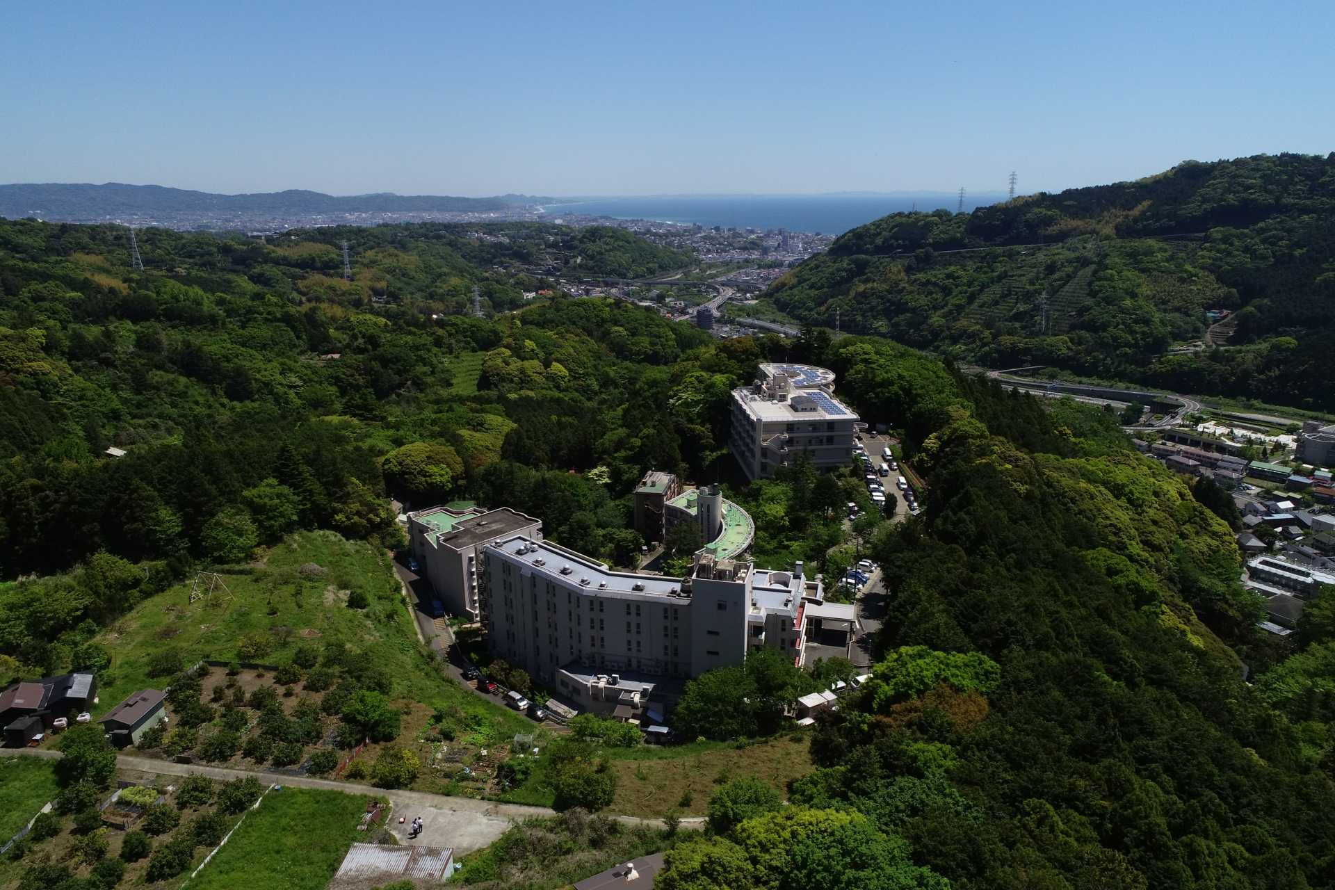 丘陵地に建つ介護施設の全景。周囲の緑地と遠景の山々が見える俯瞰写真。